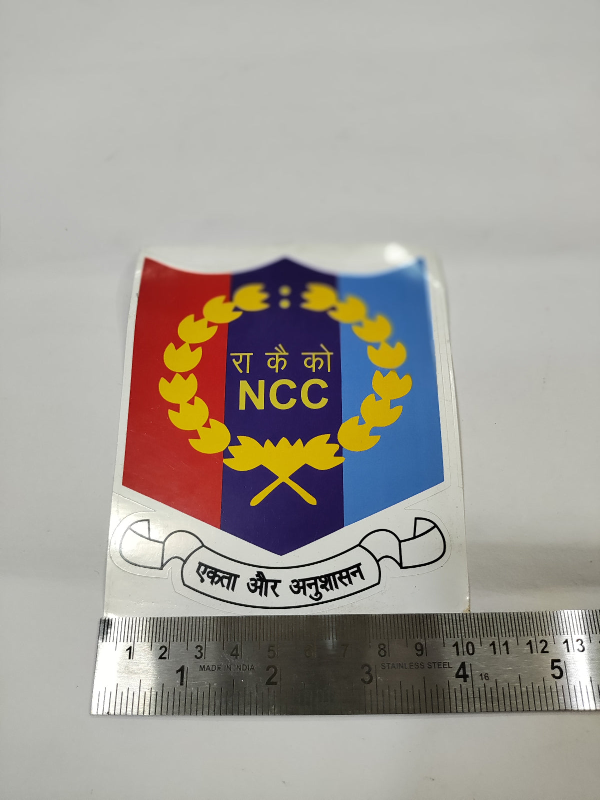NCC (National Cadet Corps) logo with a ruler for scale on a gray background